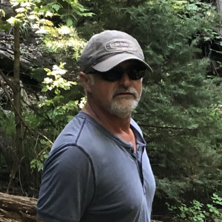 Mike Rague in the forest. He is wearing a baseball cap and dark sunglasses.