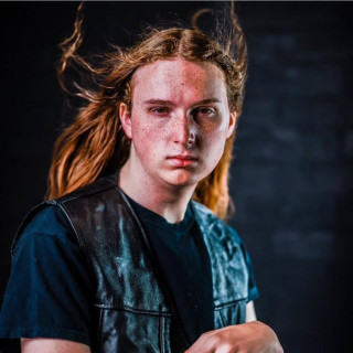 Person with long reddish brown hair, lightly blowing back with a serious facial expression. Photo credit: Hollywood Photo Studios